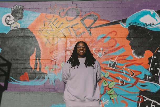 "Tree stands in front of a mural she created in Renton, with the message 'Black Lives Matter.' She is wearing a lavender sweatshirt and sweatpants."