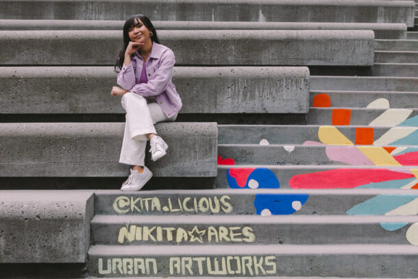 Nikita, a Filipino Female with black hair is sitting on the steps of Day One near the Spheres in South Lake Union. She is smiling with one hand under her chin and one arm resting on her knee. Beneath her you can read in chalk" @Nikita.Licious, Nikita Ares and Urban ArtWorks."