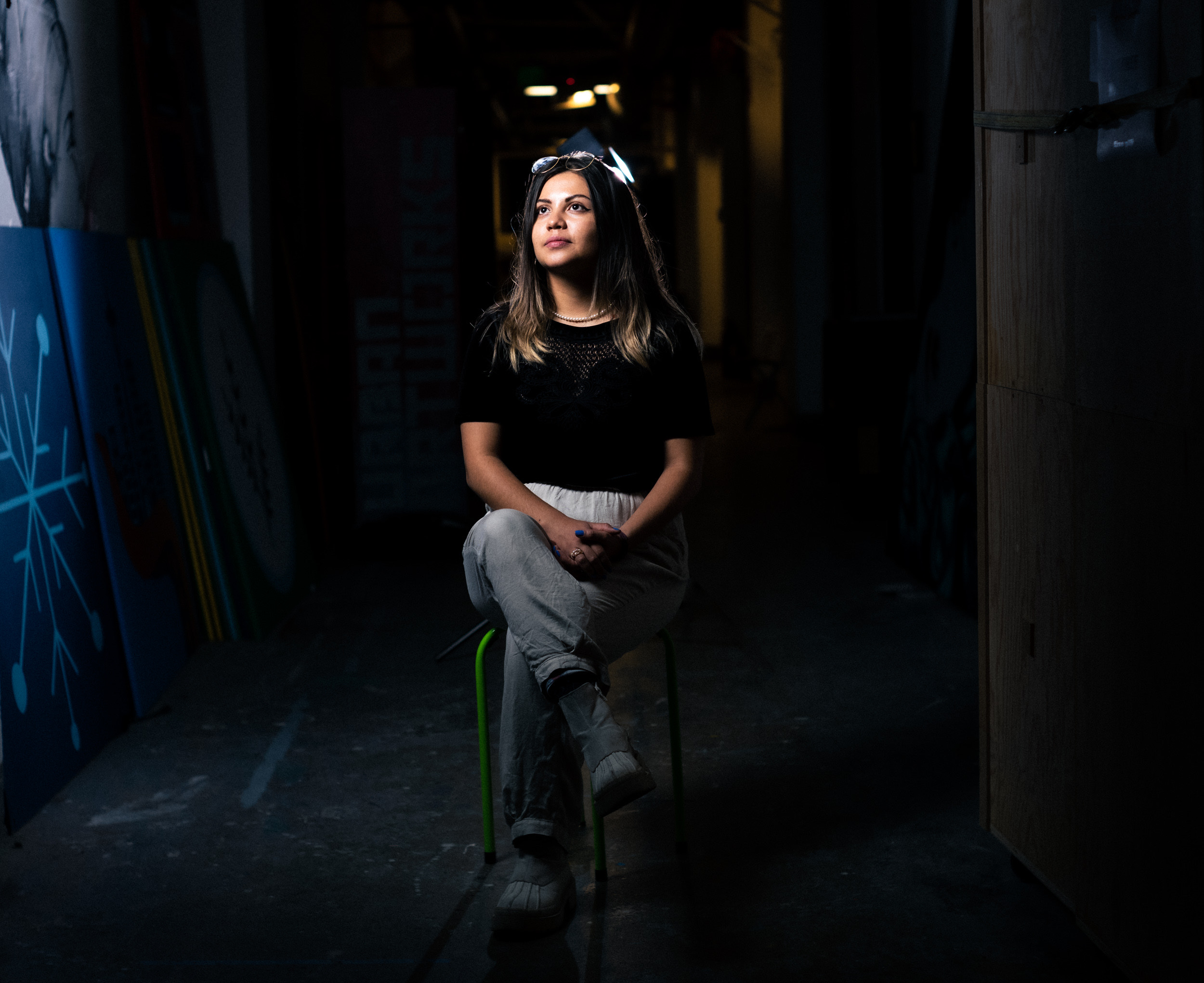 An image of Natalia Sotelo - Natalia is a Latina woman with brown eyes and currently short hair. She is wearing a black shirt with gray pants and is sitting on a stool inside of the Urban ArtWorks hallway. She is looking away from the camera
