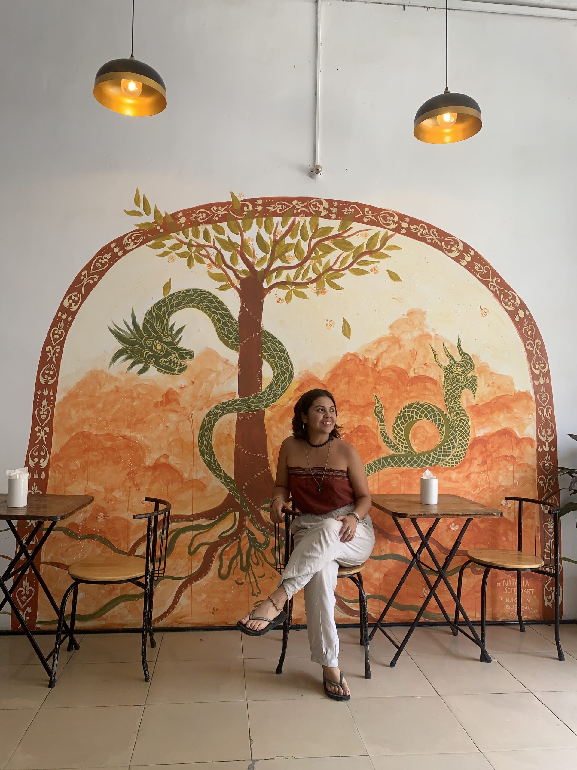 An image of Natalia Sotelo - Natalia is a Latina woman with brown eyes and currently short hair. She sits relaxedly on a chair in front of a colorful mural that she made. The mural features a vibrant scene with a green dragon coiled around a tree, set against a backdrop of warm orange tones. Natalia is wearing a strapless dark orange-colored top and white pants, accessorized with a necklaces and a bracelet. She is smiling and looking to her right.