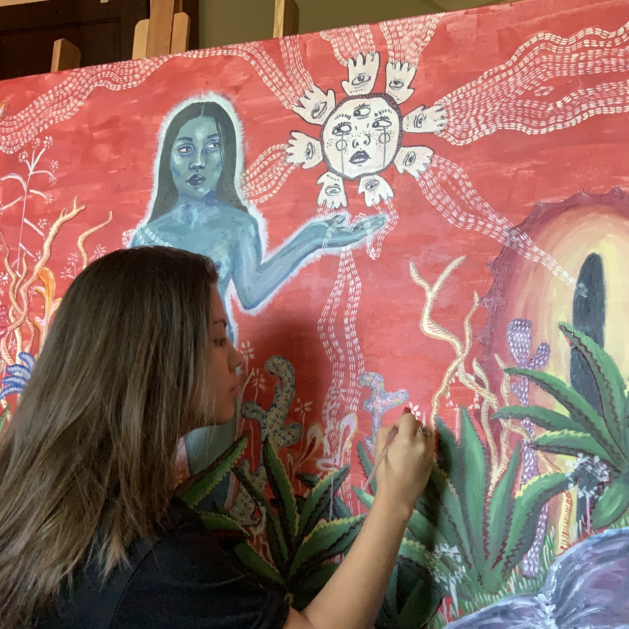 In the photo, Natalia Sotelo, a Latina woman with brown eyes and currently short hair, is intently working on a colorful painting she created. The artwork features a surreal scene with plants, a blue-skinned woman, and a sun-like figure with multiple faces and eyes, all set against a rich red background. Natalia is focused on adding details to the painting, her hand steady as she applies her brush to the canvas.