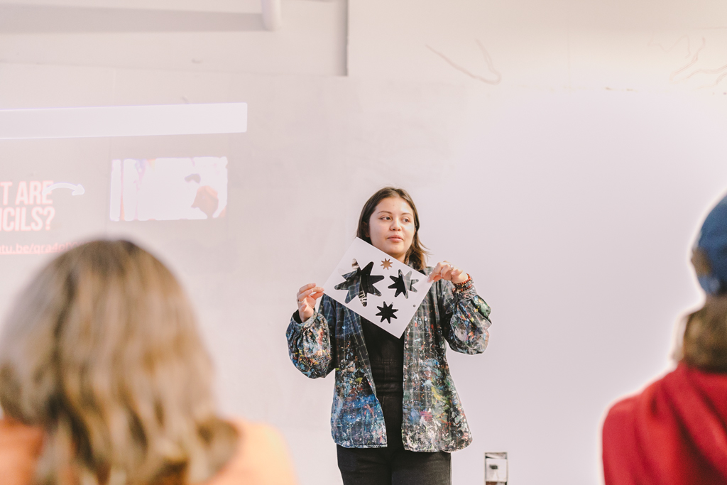 In this photo, Natalia Sotelo (she/her), a binational artist, stands at the front of a classroom holding up a design featuring several star-like shapes cut out of paper. She is teaching a group of students from Woodmoor Elementary School. Natalia is wearing a colorful, paint-splattered jacket, and the students in the foreground are focused on her demonstration. The setting is bright, with a projected image partially visible behind her, on the Urban ArtWorks studio wall.
