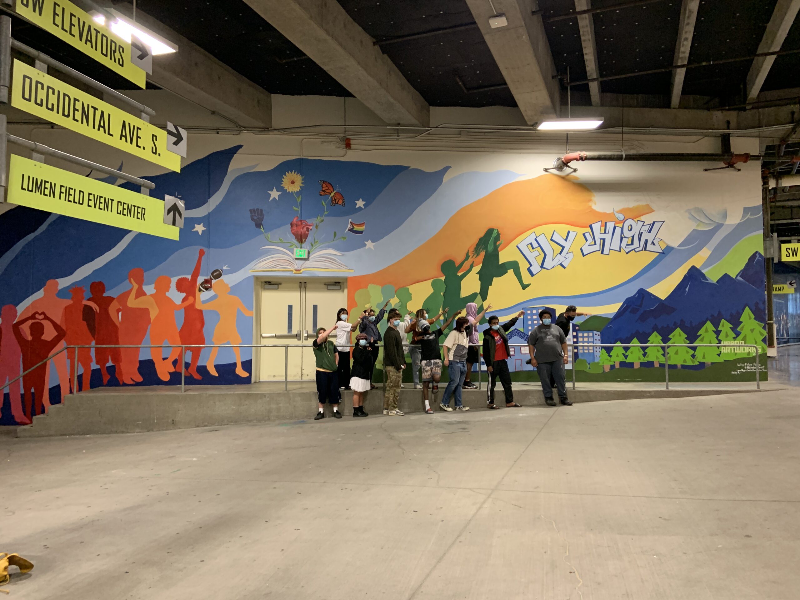The photo shows the mural at Lumen Field stadium, created through the Mural Apprentice Program in collaboration with the Seahawks. The mural features a colorful scene with various elements, including silhouettes of people in shades of red and orange on the left, and a large book with flowers, a heart, and a butterfly emerging from it. On the right side, green silhouettes of people and the words "Fly High" are set against a backdrop of mountains, trees, and buildings. Natalia and apprentices stands in front of the mural, with bright yellow signs pointing to different locations within the stadium add context to the setting.
