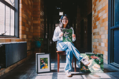 This image shows Ivy Lui, a female Chinese American artist with black hair, sitting in a hallway inside the Inscape Arts Building where Urban ArtWorks is located. She is holding one of her artworks and has other pieces displayed around her. The hallway has exposed brick walls, and natural light is coming in from a window to the left. Ivy is casually dressed in a white long-sleeve shirt and light blue jeans, with black and white sneakers. The scene captures a moment of pride and reflection as she presents her work.