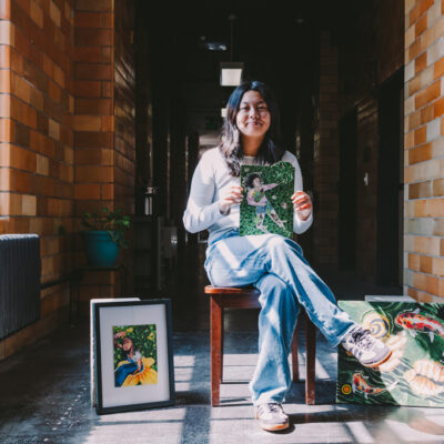 This image shows Ivy Lui, a female Chinese American artist with black hair, sitting in a hallway inside the Inscape Arts Building where Urban ArtWorks is located. She is holding one of her artworks and has other pieces displayed around her. The hallway has exposed brick walls, and natural light is coming in from a window to the left. Ivy is casually dressed in a white long-sleeve shirt and light blue jeans, with black and white sneakers. The scene captures a moment of pride and reflection as she presents her work.