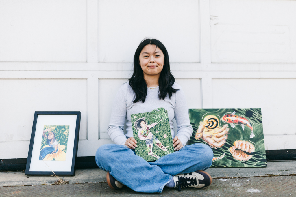This image shows Ivy Lui, a female Chinese American artist with black hair siting cross-legged in front of a white door, holding a painting in her lap. The painting depicts a woman lying on a bed of green leaves, reaching for a large, green leaf. There are two other framed paintings to her left and right. The painting on the left shows a woman standing in a field of flowers, while the painting on the right shows a hand reaching for a fish in a pond.