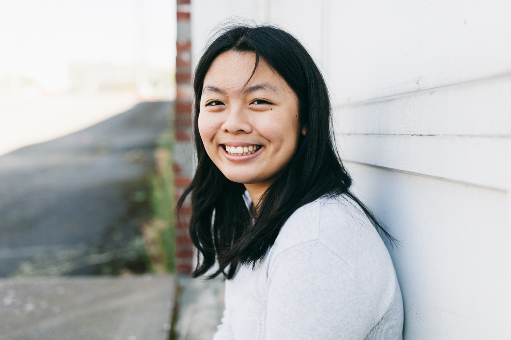 This image shows Ivy Lui, a female Chinese American artist with black hair smiling brightly at the camera. She is standing in front of a white wall with horizontal siding. The background is out of focus with some greenery.