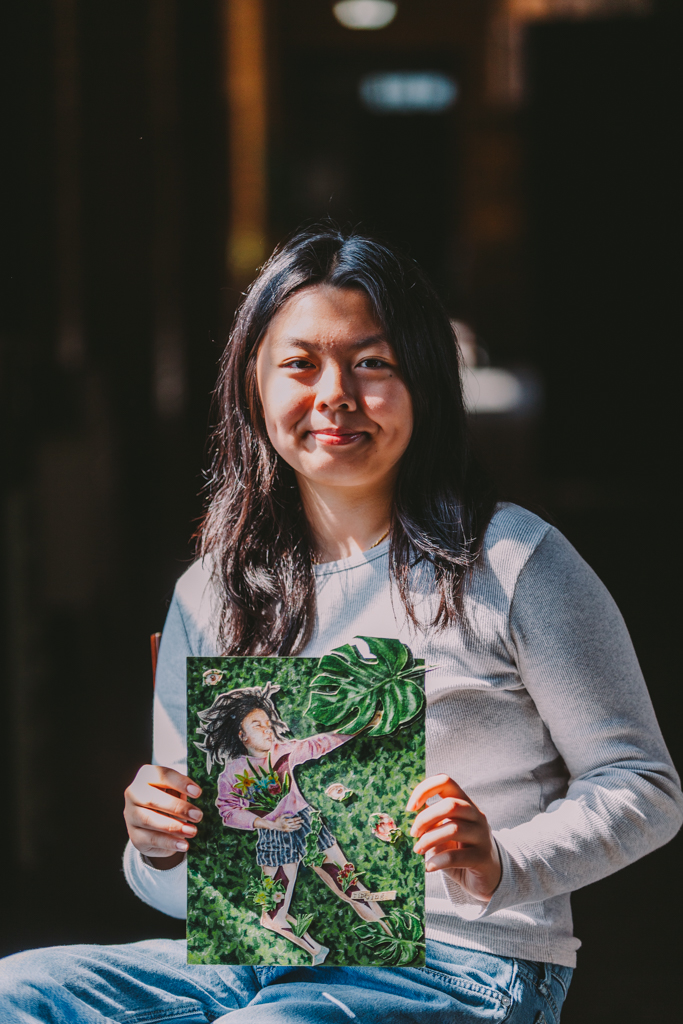 This image shows Ivy Lui, a female Chinese American artist with black hair, sitting in a hallway inside the Inscape Arts Building where Urban ArtWorks is located. She is holding one of her artworks that has a girl reaching out of the frame displayed on cut-out greenery, and she is holding pink flowers with flowers coming from cut outs on her legs.