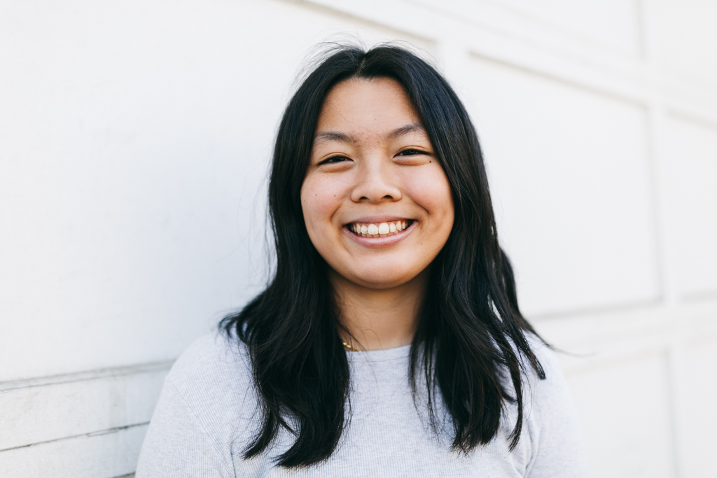 This image shows Ivy Lui, a female Chinese American artist with black hair smiling. She is facing the camera and standing in front of a white background wearing a gray long sleeve shirt.