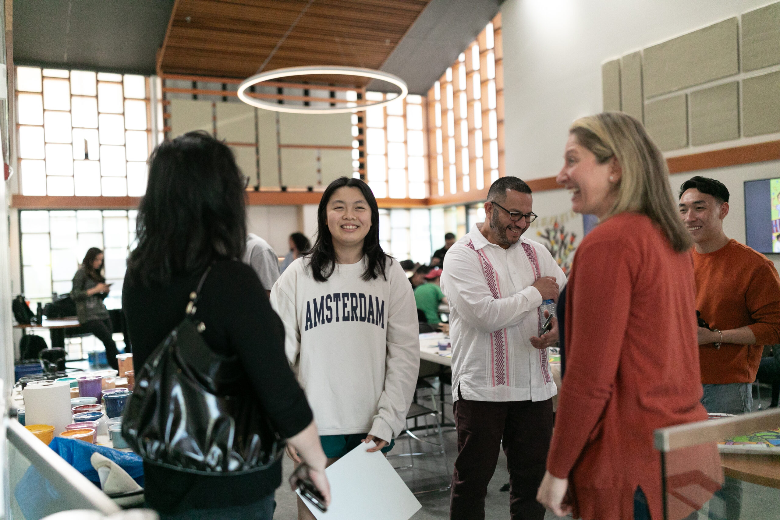 This image shows Ivy Lui, a female Chinese American artist with black hair smiling in the center of volunteers during the paint day at the Refugee Women’s Alliance (ReWA) offices. She is wearing a sweatshirt that says "Amsterdam" and she is the only person facing the camera.
