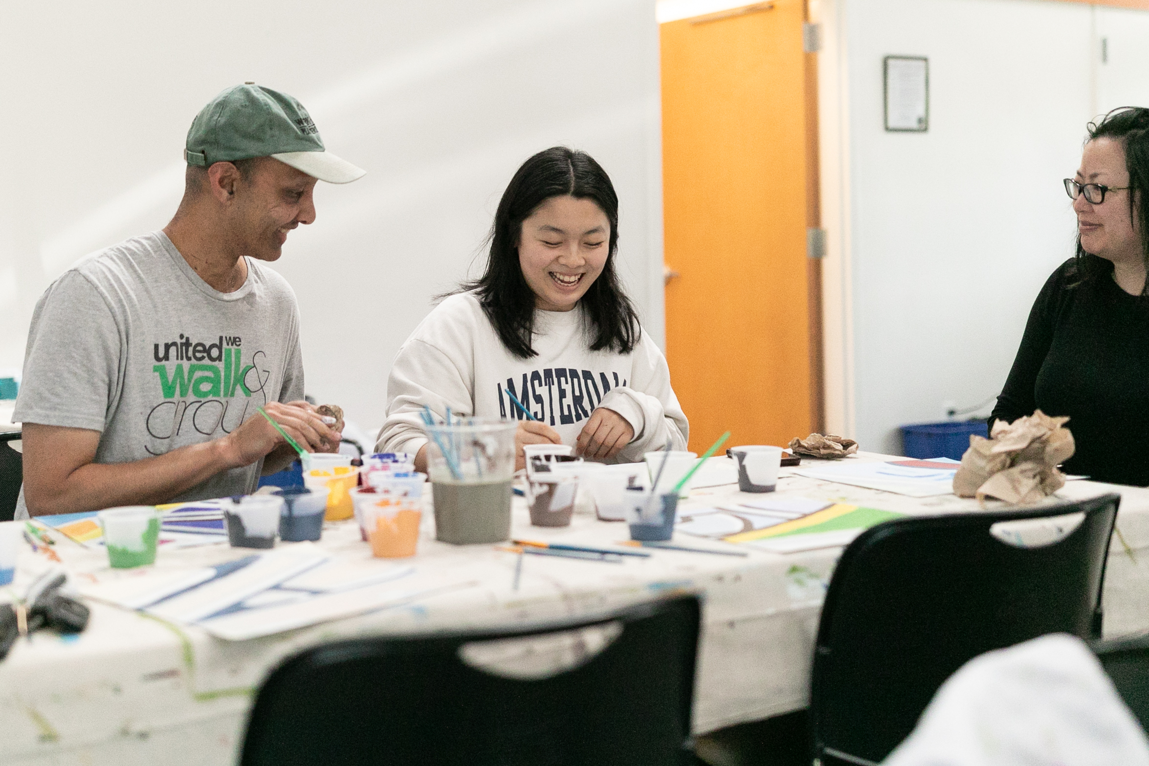 This image shows Ivy Lui, a female Chinese American artist with black hair and smiling while painting with volunteers at a table during the Refugee Women’s Alliance paint day.