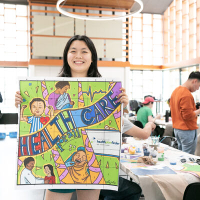 This image shows Ivy Lui, a female Chinese American artist with black hair smiling while holding her design that was chosen by the Washington Health Benefit Exchange. The design reads “Healthcare" she is standing in a room where volunteers are painting behind her, during the paint day at the Refugee Women’s Alliance (ReWA) offices.