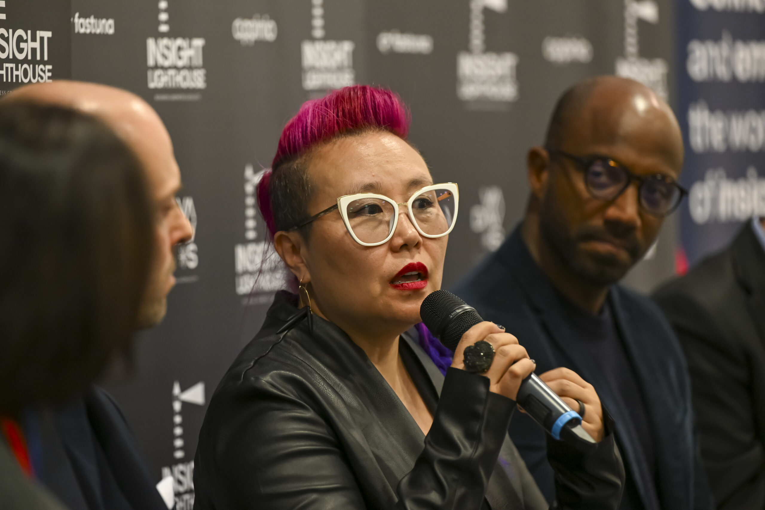 Kimfer, wearing glasses and speaking into a microphone at the Insight Lighthouse Lounge.