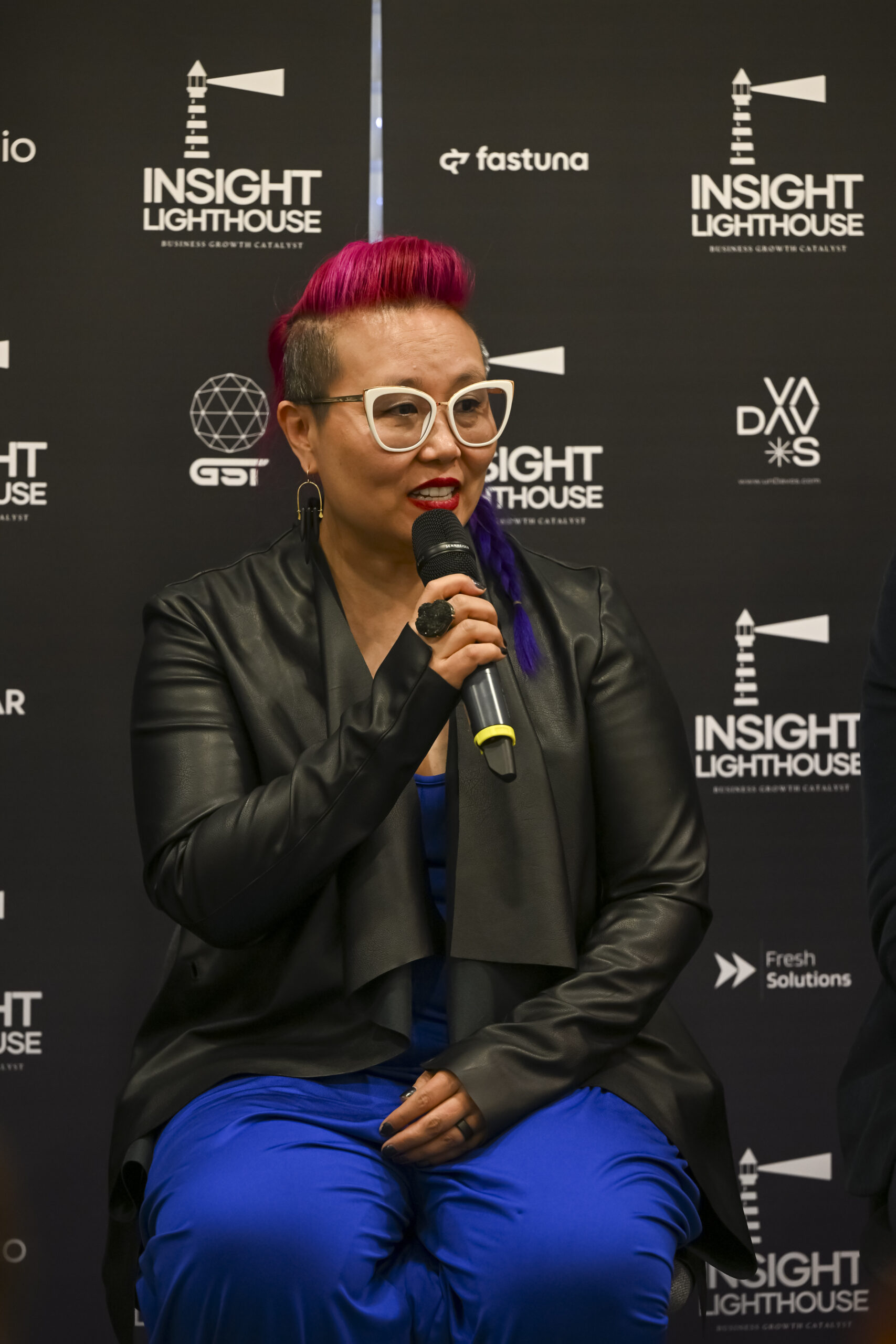 Kimfer, wearing glasses and speaking into a microphone at the Insight Lighthouse Lounge.