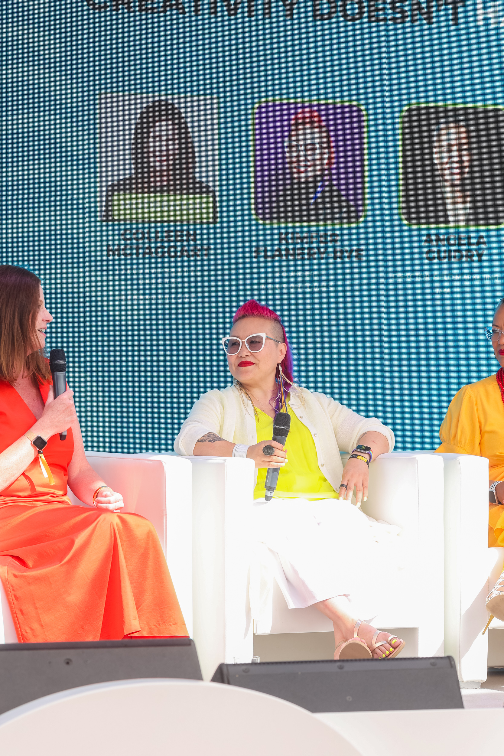 Kim "Kimfer," the Board Chair, participating in a panel discussion. Kimfer is seated in the center, holding a microphone, and dressed in a bright, summery outfit. She is wearing a neon yellow top, a white cardigan, and white pants, accessorized with white-framed glasses, a pink and purple undercut hairstyle, and colorful earrings. To her left is a panelist in an orange dress, also holding a microphone. Behind them, a large screen displays the names and photos of the panel participants, including Kimfer.