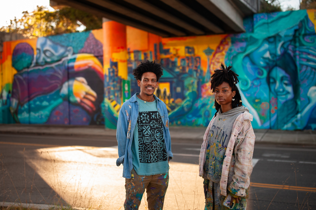 Nahom Ghirmay, a Black male with curly hair, and Jocelyn Oats are standing in front of the colorful mural in West Seattle. The mural features the Base Crew artists who placed various symbols, including images of a Base Crew Students mother, the Seattle skyline, flowers, and animals significant to First Nations culture.