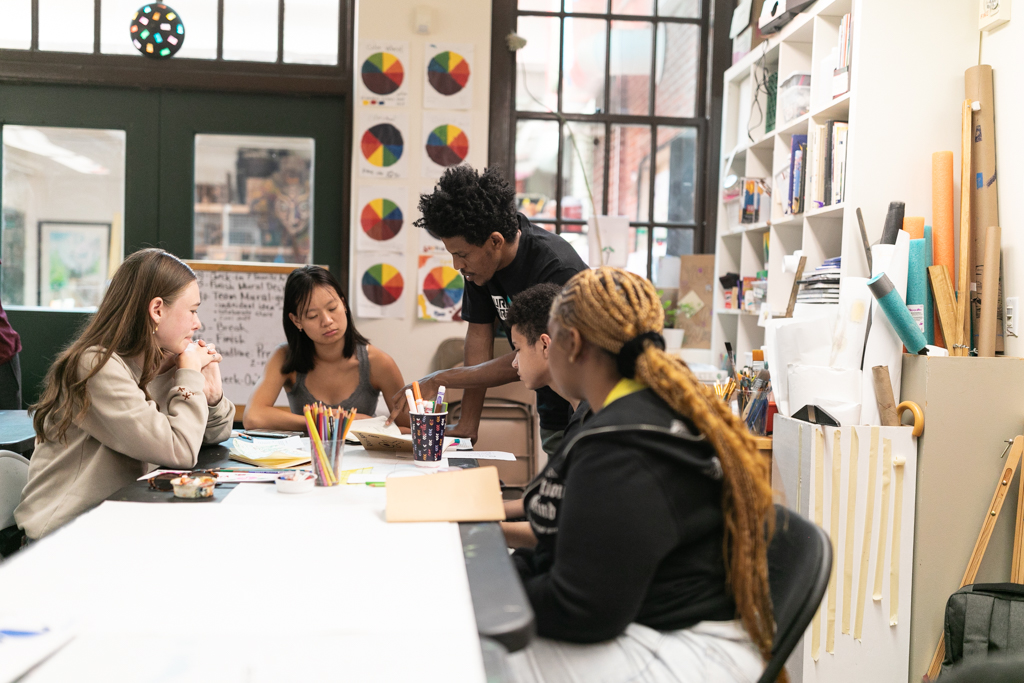 Teaching Artist Nahom Ghirmay, a Black male with brown skin and mid-length curly hair, is standing in front of the Delridge MAP students who are sitting at a table in the classroom. The environment includes a desk, whiteboard, and art on the walls.