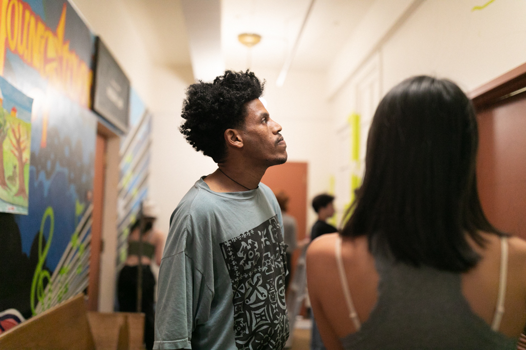 In this image, Nahom, a Black male with brown skin and mid-length curly hair, is teaching at the Delridge MAP program. He stands in a hallway with colorful murals lining the walls, focusing on the mural being created by the MAP students. Nahom wears a loose-fitting T-shirt and appears immersed in observing the artwork. Several students surround him, including one just in front of him with their back to the camera.