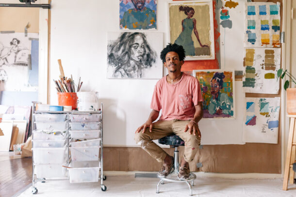 Teaching Artist Nahom, a Black male with brown skin and mid-length curly hair, is seated on a stool in his art studio. He is wearing a muted pink T-shirt and tan pants with paint stains. Nahom sits relaxed, with his hands resting on his knees. The studio is bright and filled with Nahom's artwork displayed on the wall behind him. There are various portrait paintings and studies, each showing different styles and color palettes. To his left, a cart with drawers holds an assortment of art supplies.