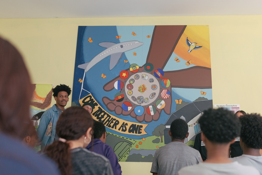 The image shows the classroom in the SeaTac MAP final celebration, where Teaching Artist Nahom, a Black male with brown skin and mid-length curly hair, stands in front of the mural —the finished product of a collaborative project. Around him are students, visible from the backs of their heads.