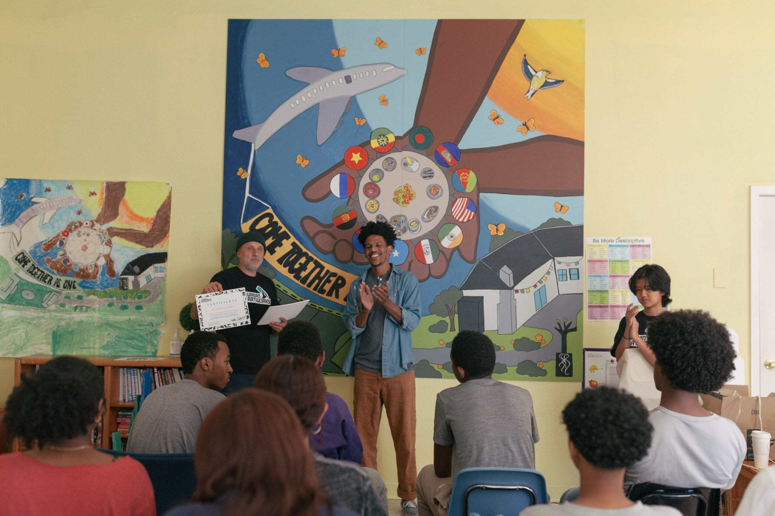The image captures a classroom scene from the SeaTac MAP final celebration, where Teaching Artist Nahom, a Black male with brown skin and mid-length curly hair, is teaching alongside Teaching Artist Seth. Nahom stands near a vibrant mural on the wall behind him—the finished product of a collaborative project. To the left of the mural, a drawing on the wall shows the original sketch that inspired the mural, highlighting the progression from concept to completion. Nahom is clapping and smiling to celebrate the students achievements.