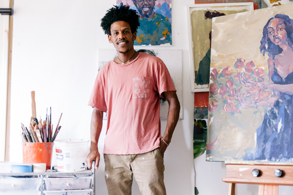 This image shows Teaching Artist Nahom, a Black male with brown skin and mid-length curly hair, in his art studio. He is wearing a muted pink T-shirt and tan pants with paint stains. Nahom stands next to his artwork relaxed with his right hand in his pants pocket. There are various portrait paintings and studies, each showing different styles and color palettes. To his left, a cart with drawers holds an assortment of art supplies. The painting on the easel shows a woman with long hair in a dress holding flowers.