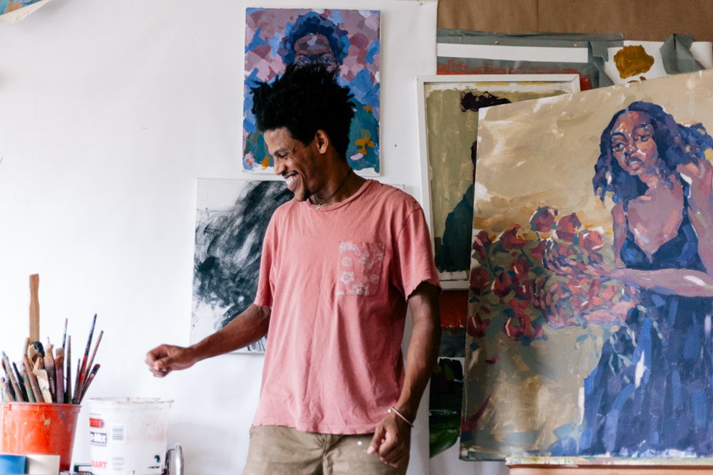 In this image, Teaching Artist Nahom, a Black male with brown skin and mid-length curly hair, is in his art studio, Nahom's artwork is displayed on the easel next him. To his left, a cart with drawers holds an assortment of art supplies, including brushes. Nahom is reaching for a brush while smiling.