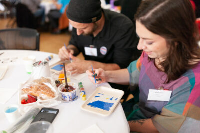 Two people sit at a round table painting small canvases, surrounded by art supplies and a tray of breakfast items, including a croissant and fruit. Both are focused on their art, which were parts of designs made by Base Crew artists.