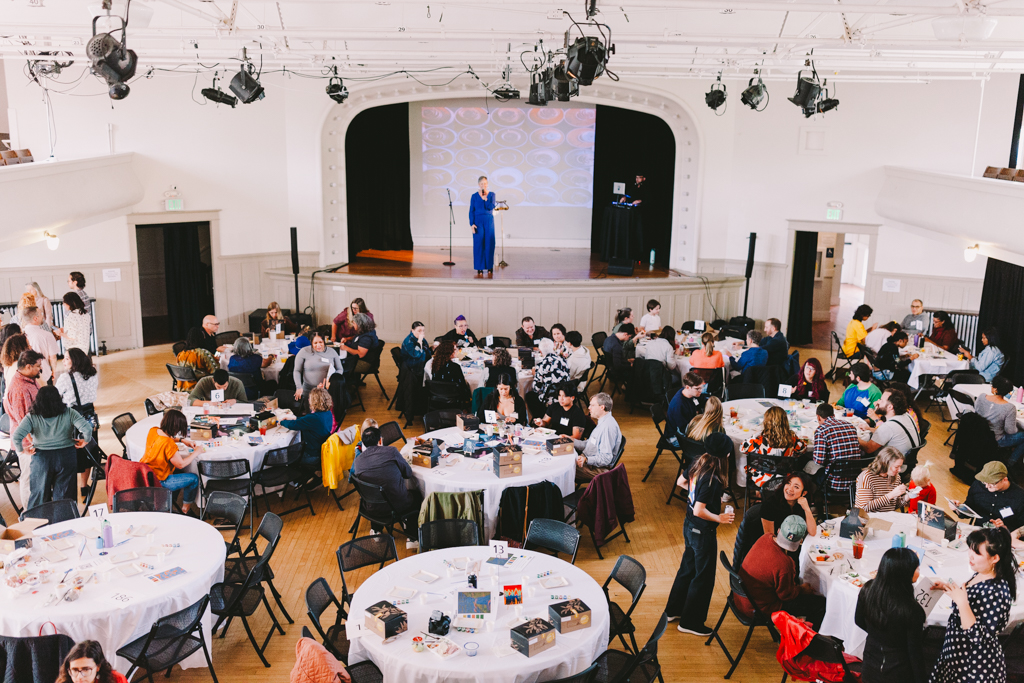 a view from the third floor inside of Washington Hall, Brush and Brunch is happening below. There are tables of people bidding on auction items with then auctioneer standing on stage in a blue jumpsuit.