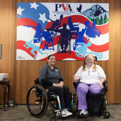 Two people in mobility devices in a LiftZone sponsored by Comcast at the Paralyzed Veterans of America (PVA) Northwest Chapter. Behind them is the mural he created by Charles Connor for PVA.
