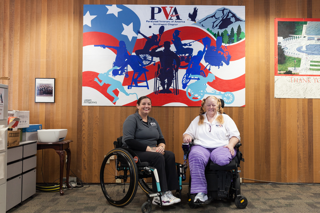 Two people in mobility devices in a LiftZone sponsored by Comcast at the Paralyzed Veterans of America (PVA) Northwest Chapter. Behind them is the mural he created by Charles Connor for PVA.