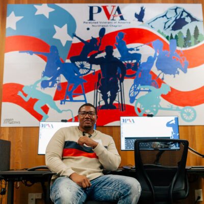 The artist Charles Connor sitting in a LiftZone sponsored by Comcast at the Paralyzed Veterans of America (PVA) Northwest Chapter. He is wearing glasses, a beige sweater with red and navy blue stripes, and distressed jeans. Behind him is the mural he created for PVA with a patriotic theme, incorporating red and white stripes, blue silhouettes of individuals in different mobility devices in various activities like sports, and a mountainous landscape in the background. The mural includes the text "PVA" for Paralyzed Veterans of America, Northwest Chapter, and the Urban ArtWorks logo.