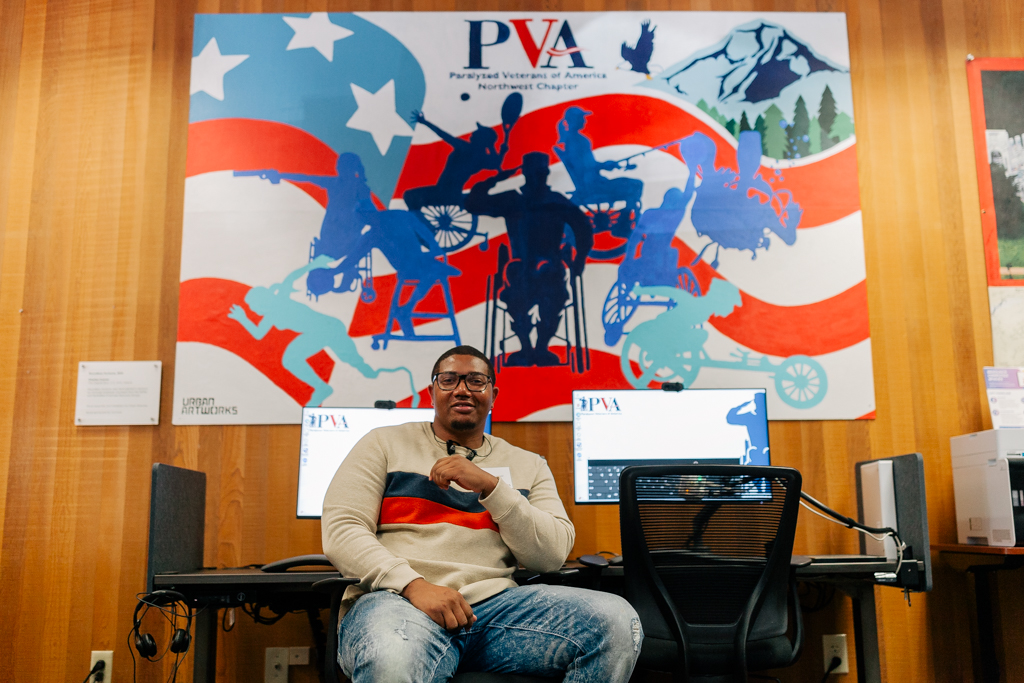 The artist Charles Connor sitting in a LiftZone sponsored by Comcast at the Paralyzed Veterans of America (PVA) Northwest Chapter. He is wearing glasses, a beige sweater with red and navy blue stripes, and distressed jeans. Behind him is the mural he created for PVA with a patriotic theme, incorporating red and white stripes, blue silhouettes of individuals in different mobility devices in various activities like sports, and a mountainous landscape in the background. The mural includes the text "PVA" for Paralyzed Veterans of America, Northwest Chapter, and the Urban ArtWorks logo.