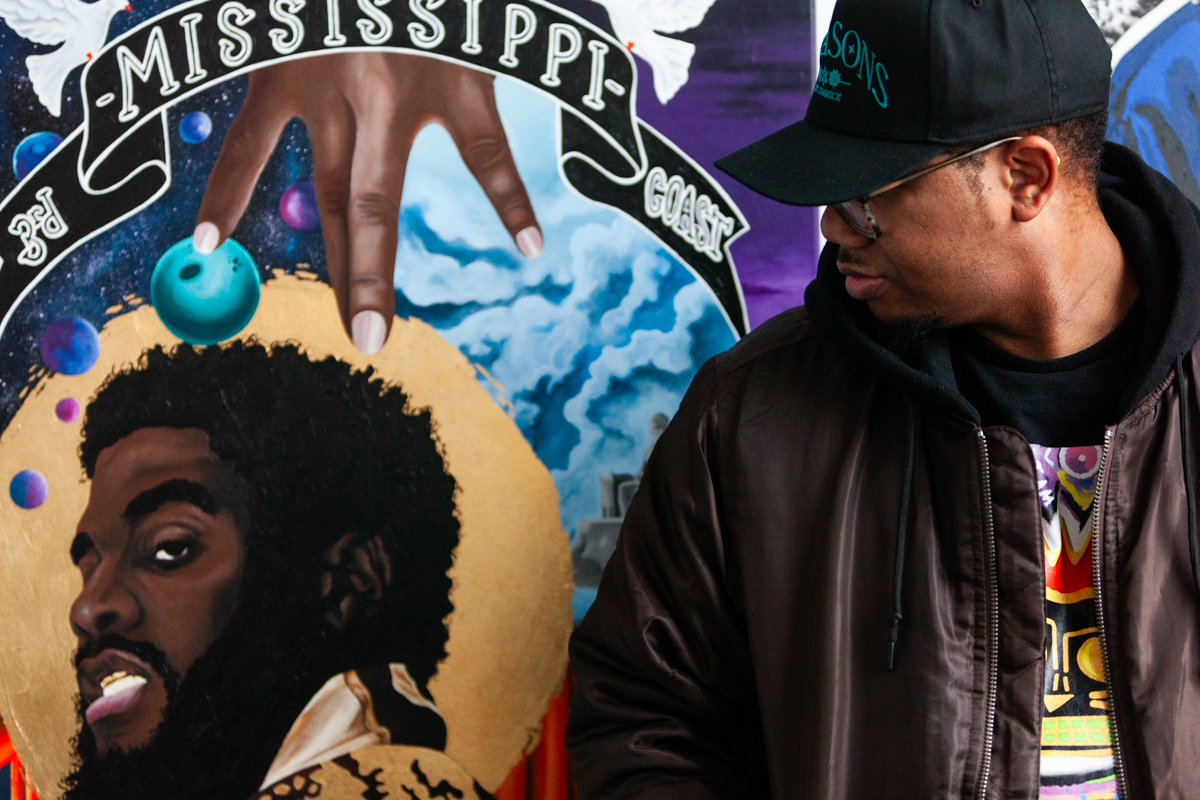 The artist Charles Connor wearing a black cap, glasses, and a dark brown bomber jacket with a black hoodie underneath. Behind him, there are three or his paintings with a mix of realism and graphic elements along with mixed media’s like fabric and texture. The mural includes a detailed portrait of K.R.I.T., a rapper and record producer. A banner that reads "Mississippi Coast."