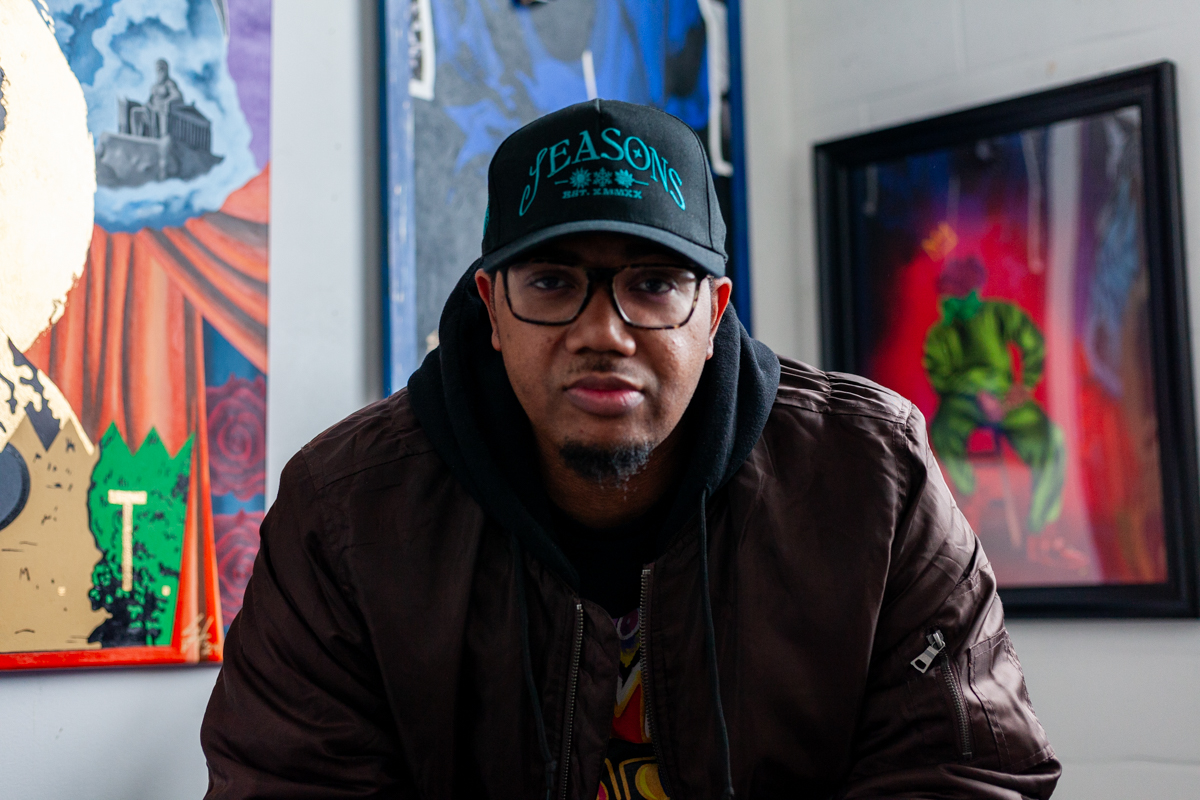 The artist Charles Connor wearing a black cap, glasses, and a dark brown bomber jacket with a black hoodie underneath. Behind him, there are three or his paintings with a mix of realism and graphic elements along with mixed media’s like fabric and texture.