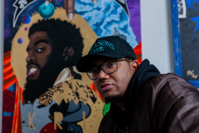 The artist Charles Connor wearing a black cap, glasses, and a dark brown bomber jacket with a black hoodie underneath. Behind him, there are three or his paintings with a mix of realism and graphic elements along with mixed media’s like fabric and texture. The mural includes a detailed portrait of K.R.I.T., a rapper and record producer. A banner that reads "Mississippi Coast."