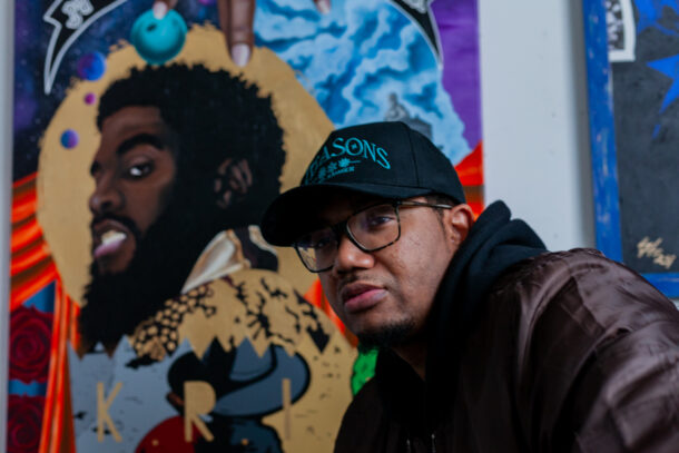 The artist Charles Connor wearing a black cap, glasses, and a dark brown bomber jacket with a black hoodie underneath. Behind him, there are three or his paintings with a mix of realism and graphic elements along with mixed media’s like fabric and texture. The mural includes a detailed portrait of K.R.I.T., a rapper and record producer. A banner that reads "Mississippi Coast."