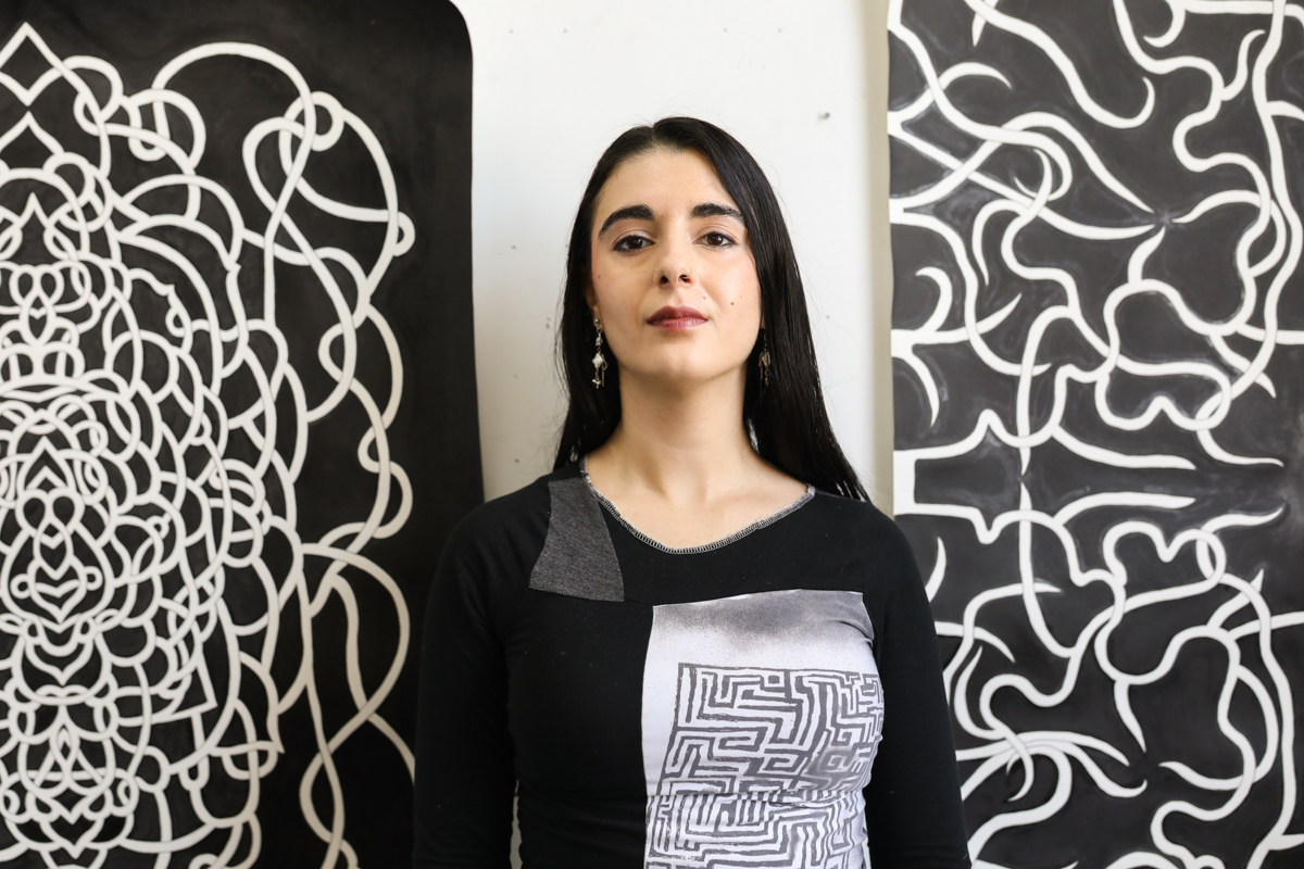 Ani standing between her two large black and white detailed artworks. Ani has dark brown/black hair, dark brown eyes, and pale olive skin. She wears a black top with a gray patch and a long denim skirt.