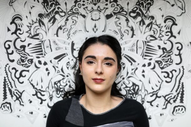 Ani against her highly detailed black and white large painting. Ani has dark brown/black hair, and dark brown eyes and pale olive skin. She wears a black top with a patch of gray near the neckline, and the centered artwork Ani created displayed behind her almost forms a frame, enhancing the visual impact of the portrait.