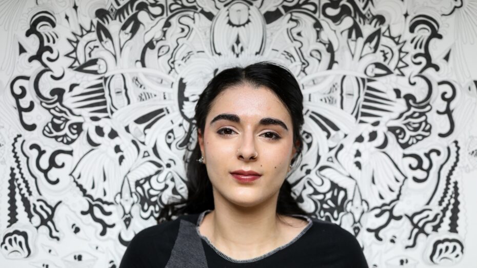Ani against her highly detailed black and white large painting. Ani has dark brown/black hair, and dark brown eyes and pale olive skin. She wears a black top with a patch of gray near the neckline, and the centered artwork Ani created displayed behind her almost forms a frame, enhancing the visual impact of the portrait.