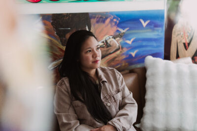 Shayla, a Filipina-American woman with medium brown skin, long black hair, and round-almond brown eyes, smiles while looking away from the camera with her artwork hanging in the background of her studio. She is sitting on a couching.