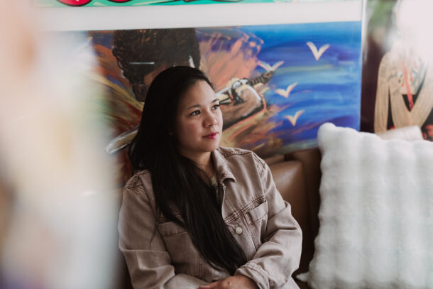 Shayla, a Filipina-American woman with medium brown skin, long black hair, and round-almond brown eyes, smiles while looking away from the camera with her artwork hanging in the background of her studio. She is sitting on a couching.