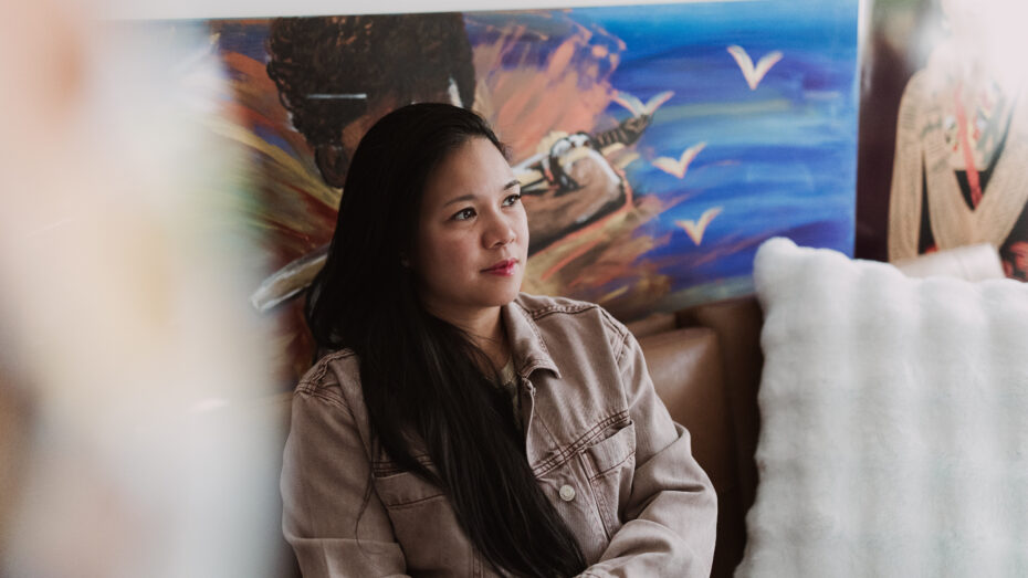 Shayla, a Filipina-American woman with medium brown skin, long black hair, and round-almond brown eyes, smiles while looking away from the camera with her artwork hanging in the background of her studio. She is sitting on a couching.
