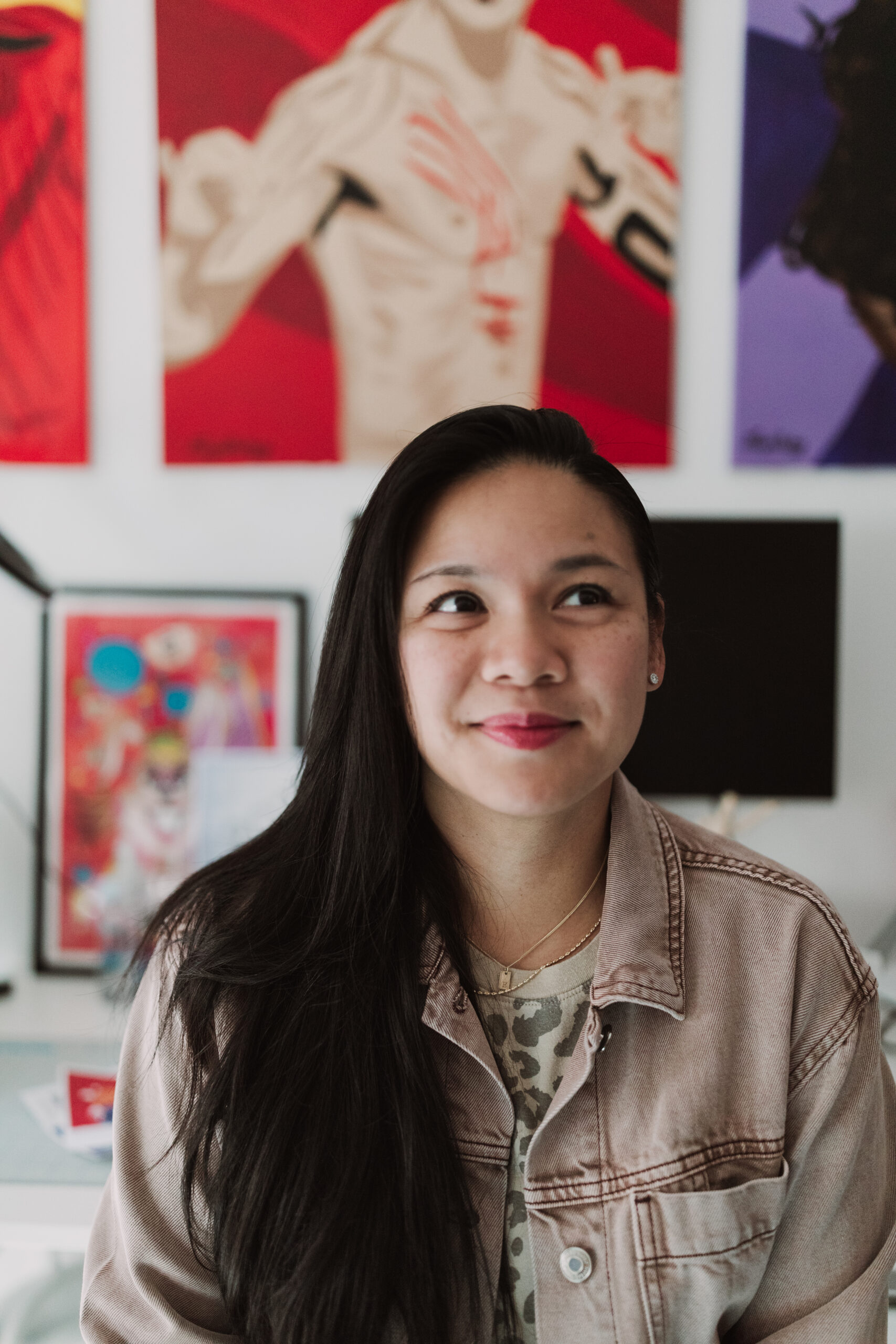 Shayla, a Filipina-American woman with medium brown skin, long black hair, and round-almond brown eyes, smiles while looking away from the camera with her artwork hanging in the background of her studio.
