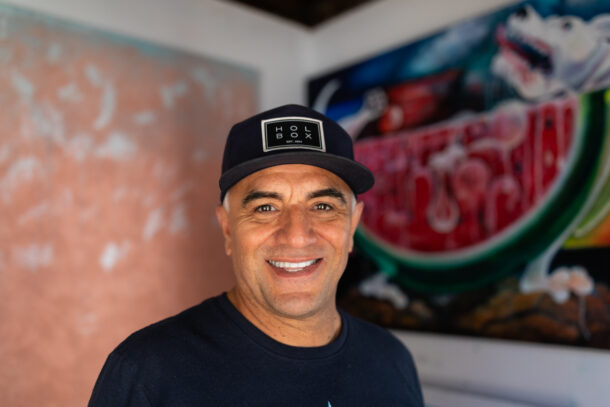 Felipe standing in his studio, smiling warmly up at the camera. Behind him, painting in progress including a bright watermelon scene to his right and a soft orange abstraction painting to his left.