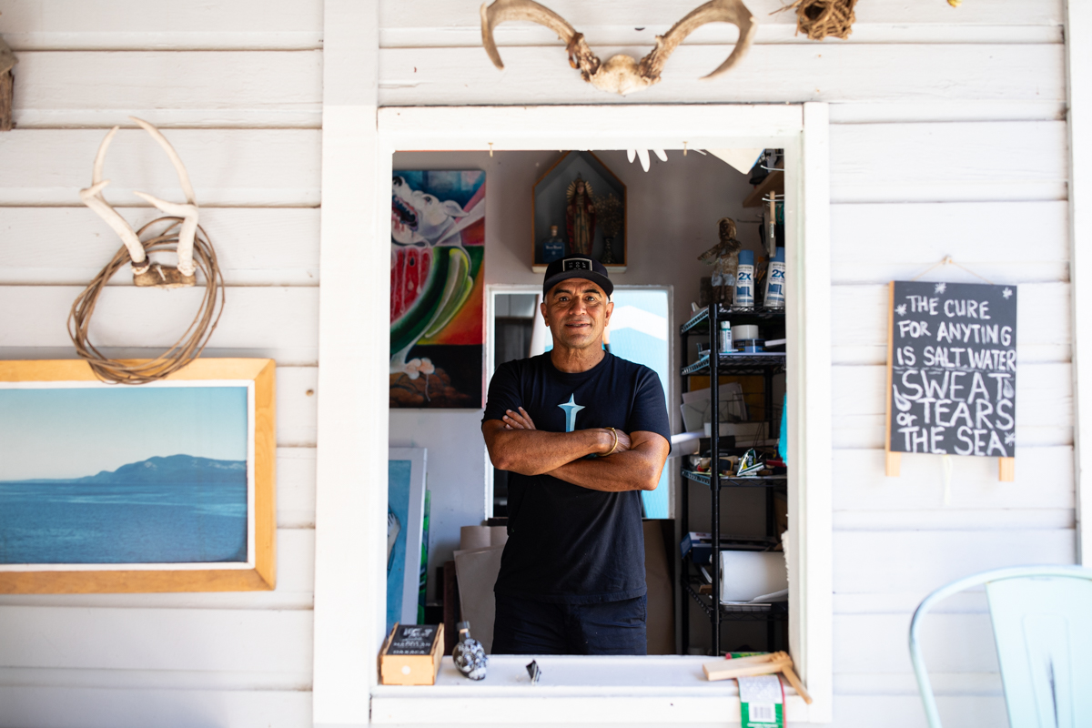 Felipe stands inside his studio surrounded by paintings in progress, seen through a window framed by antlers and art pieces hanging outside. Felipe is standing with his arms crossed.