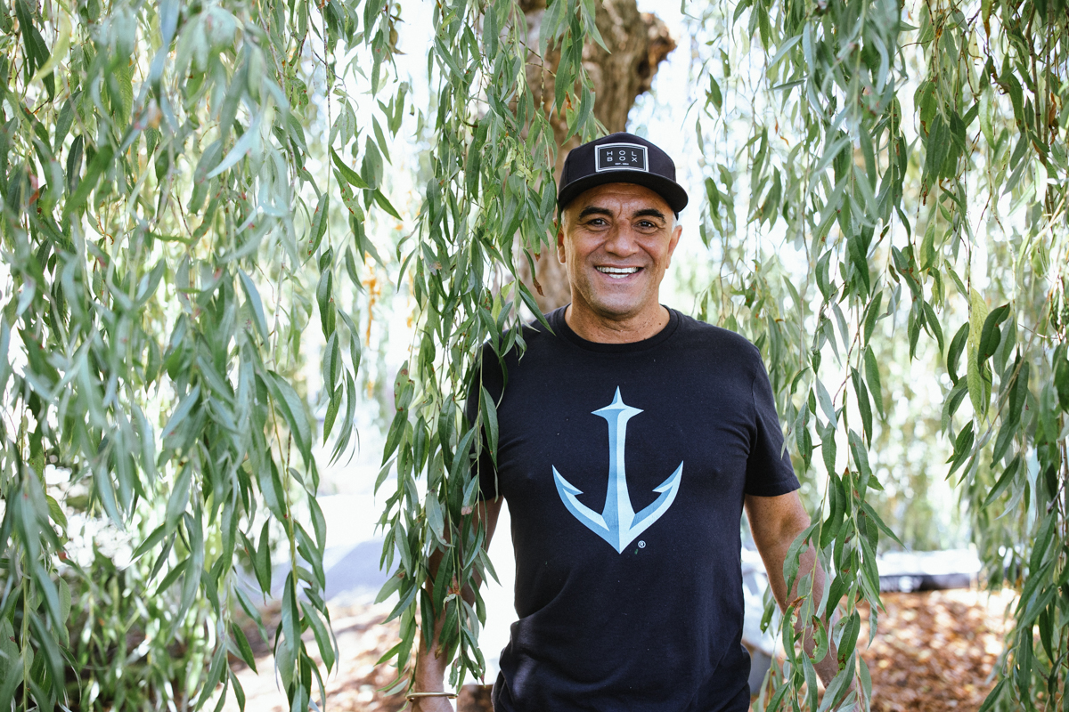 Felipe stands beneath a large weeping willow, its sweeping branches filling most of the frame surround him with cascading greenery.