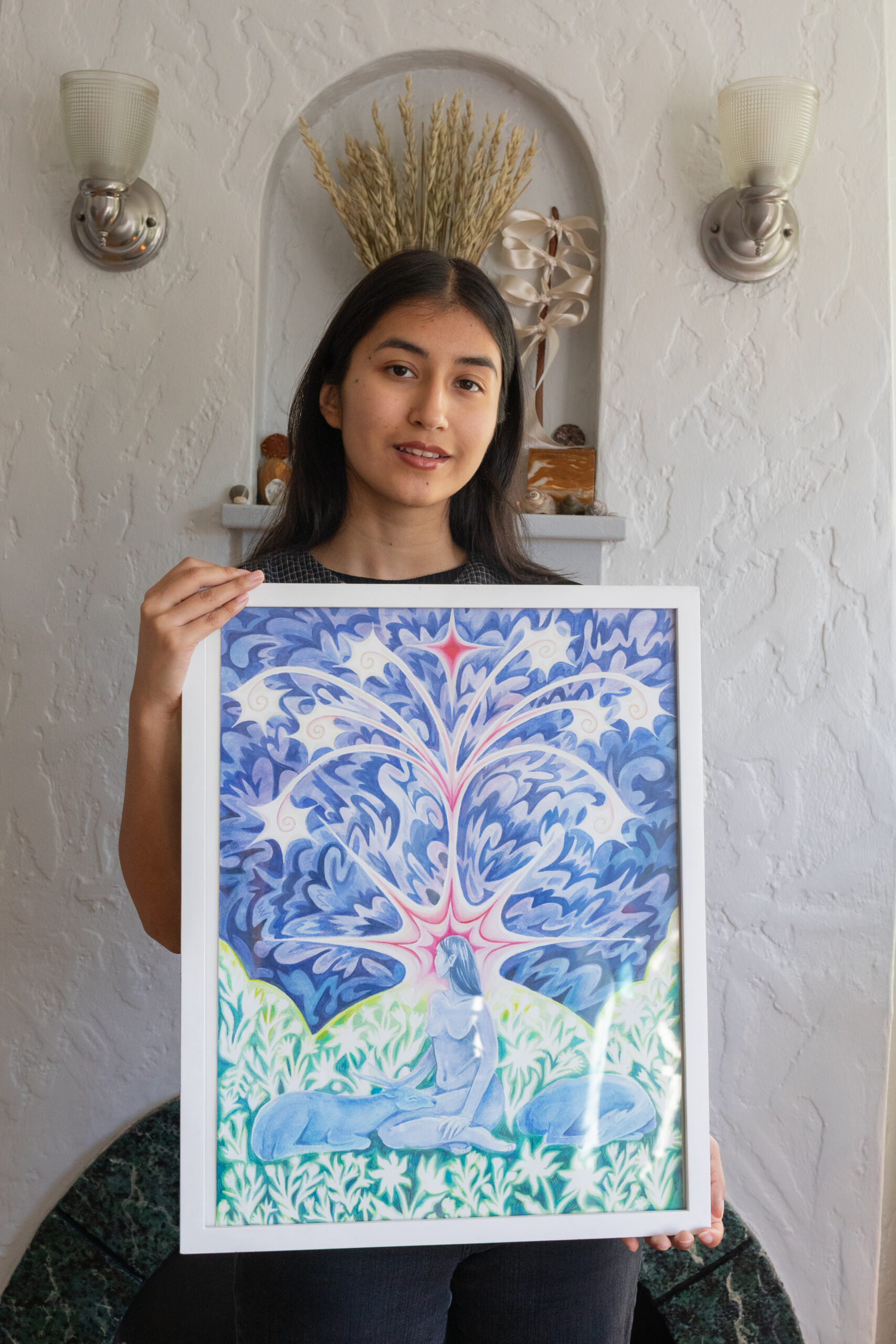 Grace woman with dark hair and tan skin smiling is smiling while holding her framed artwork in her studio.