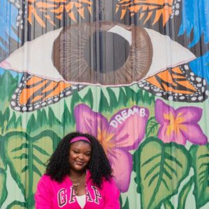 Dream Bernard sitting in front of her Mural, the mural has a brown colored eye with monarch butterfly wings and flowers.