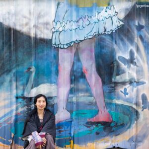Emma Zhao sitting in front of her mural. The mural has a person wearing a skirt standing on water. There are two Swans and butterflies surrounding with the moon behind them.